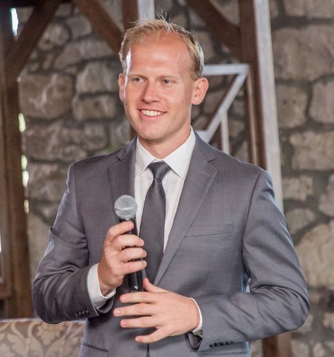 Photo of Patrick Jachyra smiling while wearing a grey suit and tie and holding a microphone.