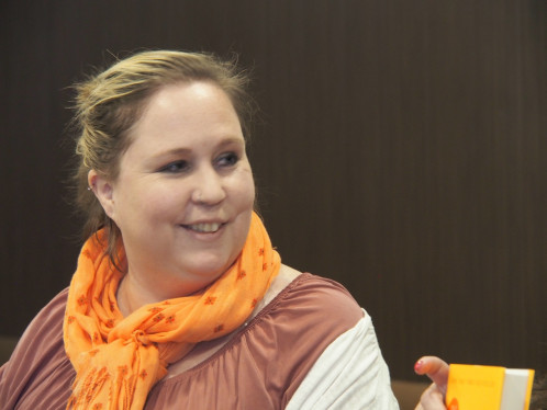 A photo of webinar host Stephanie Moeser, a smiling woman with hair tied back, wearing a brown and white shirt and an orange scarf