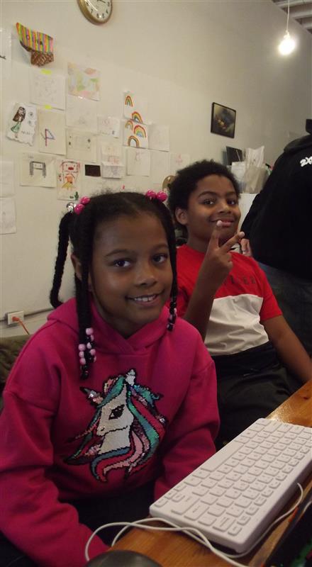 Child in bright pink hoodie smiles at the camera. In the background, another child in a red and white shirt makes a peace sign and smiles at the camera.