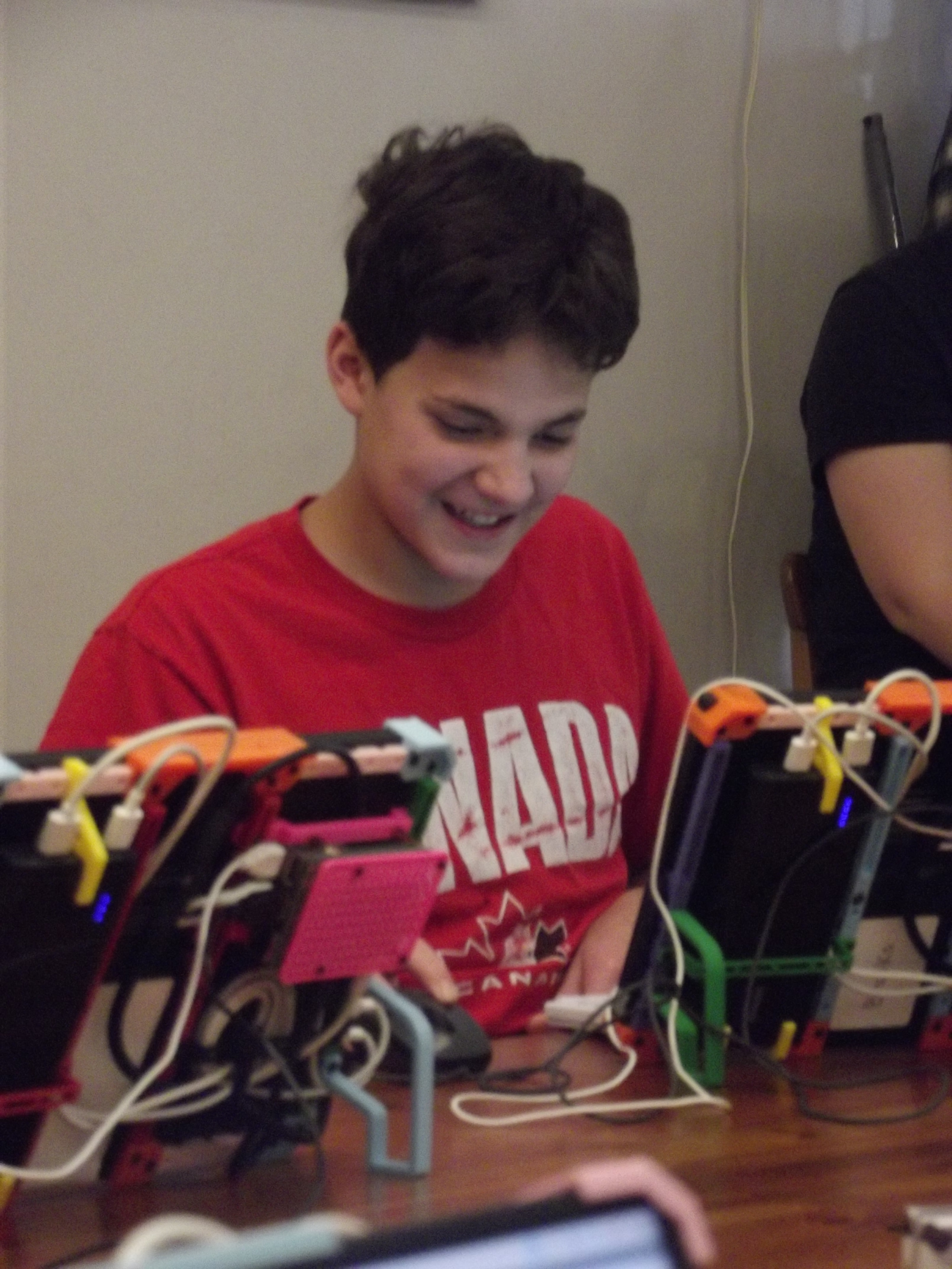 Child in red shirt smiles while looking at 3D printing devices in front of him. 