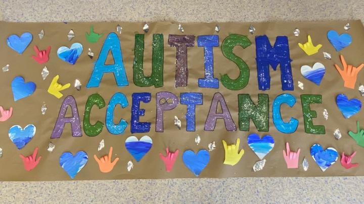 A brown banner with the words Autism Acceptance in blue, green, and purple block letters with blue hearts and multicoloured hands glued around it.