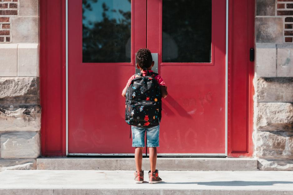 A child wearing a backpack waits outside red school doors