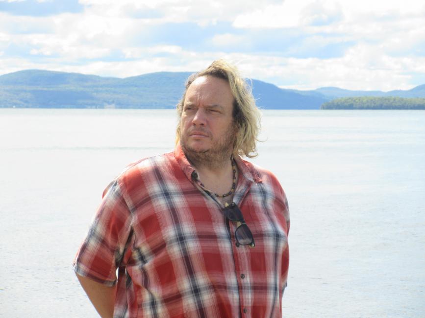 Carl Parker, with chin length hair and wearing a plaid shirt, gazes wistfully into the distance. Behind him is a lake