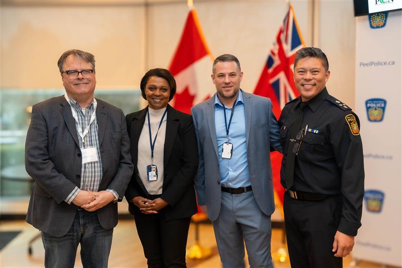 Four people stand while smiling in front of the Canadian flag. 