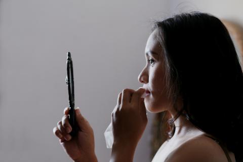 A woman looking in a handheld mirror while wiping off lipstick