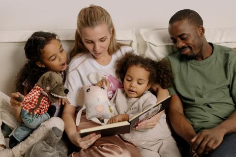 Family of four people, two daughters, a mother and a father reading a book together on the couch