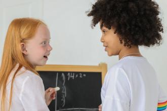 One girl holding chalk teaches another girl math in front of a mini chalkboard